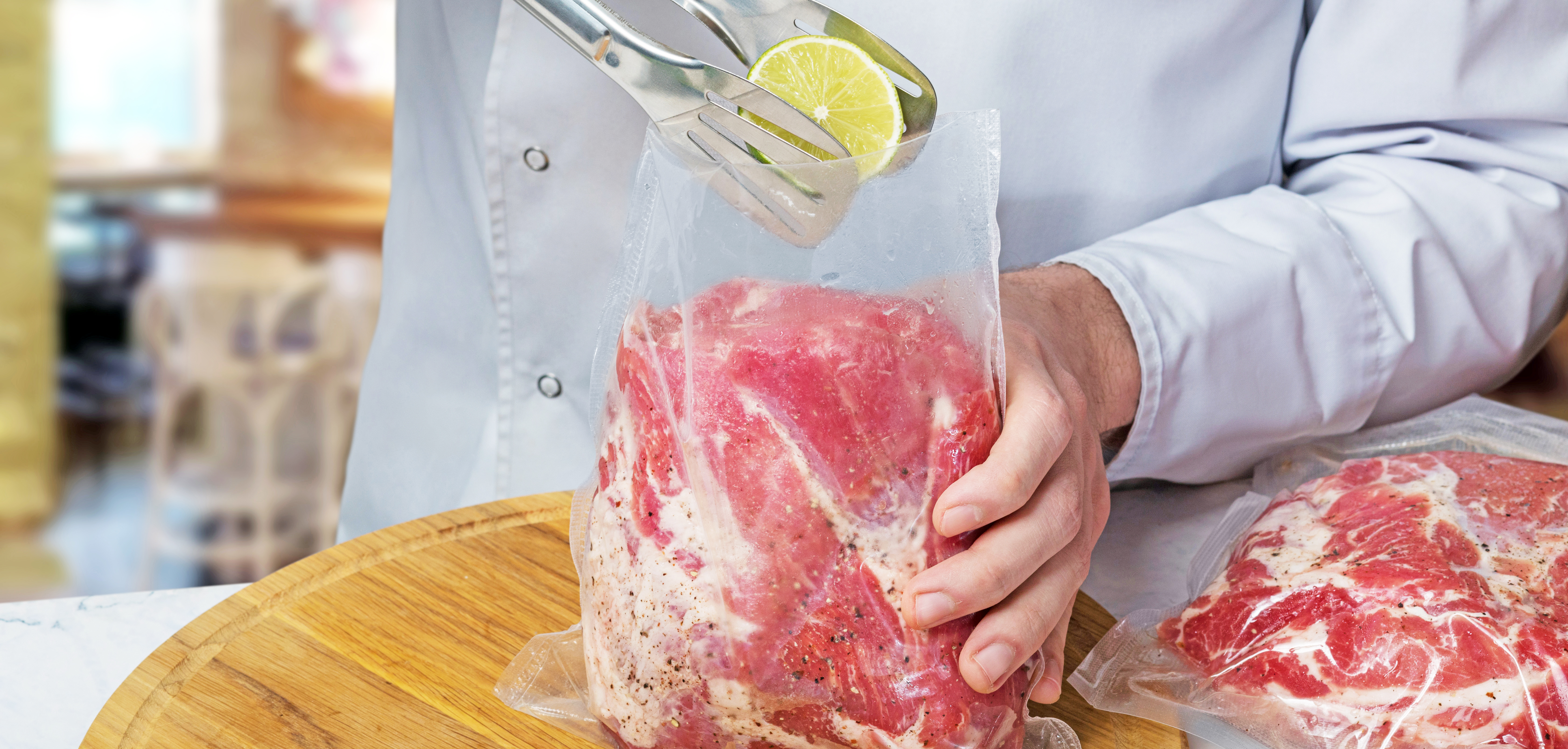 Image of a chef in a white coat using tongs to put a lemon into a vacuum packaging pouch filled with raw meat